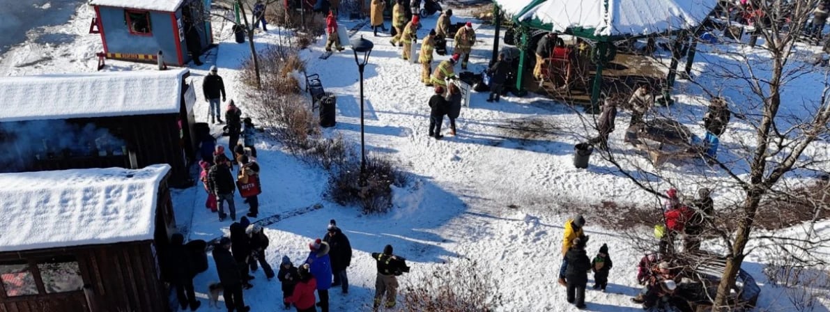 Des familles et des pompiers autour des cabanes et du kiosque enneigés lors du Noël magique